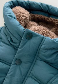 Türkise Puffjacke mit einem Kragen aus einem flauschigen, hellbraunen Futter, zwei dunklen Knöpfen und einer gesteppten Textur. Der Stoff hat ein glattes Finish.