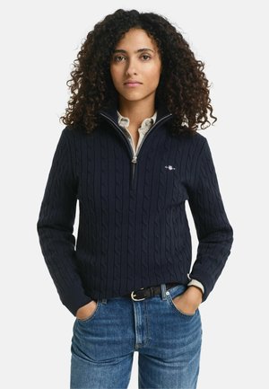 Navy cable-knit pullover with a high collar, featuring a zipper and small logo on the chest. Worn over a light-colored shirt.