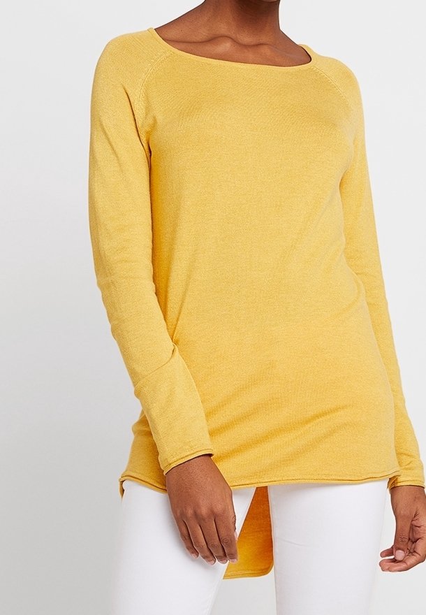 Femme portant une tunique jaune à manches longues avec un ourlet asymétrique sur un pantalon blanc, debout devant un fond clair uni.
