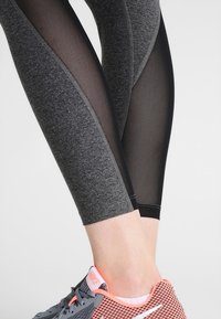 Leggings grigi con pannelli in rete e dettagli neri, caratterizzati da un design aderente. Abbinati a una scarpa da ginnastica grigia e rosa.