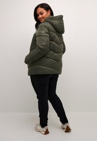 Olijfgroene pufferjack met capuchon, voorzien van een quiltpatroon en elastische details op de mouwen. Gecombineerd met zwarte broek en witte sneakers.