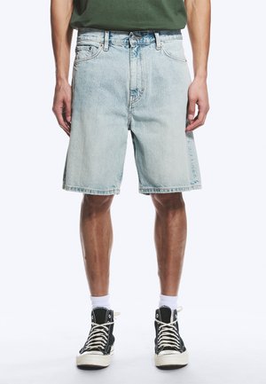 Homme portant un short en jean bleu clair, des chaussettes blanches et des baskets montantes noires, debout avec les mains détendues le long du corps.
