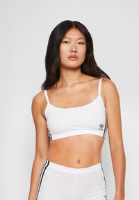 Mulher com cabelo escuro e longo, vestindo um top cropped branco da Adidas e calças combinando com riscas pretas, de pé contra um fundo simples.