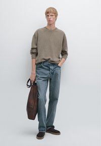 Bruine gebreide trui, lichtblauwe jeans, bruine leren schoudertas en donkere schoenen. Model staat tegen een witte achtergrond.