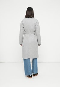 Persona in piedi girata di spalle, che indossa un cappotto grigio chiaro con cintura, jeans a gamba larga blu e sneakers nere con suola marrone, su sfondo bianco.