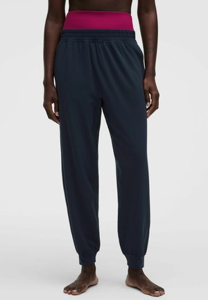 Persoon draagt losse marineblauwe joggingbroek met elastische taille en boorden, gecombineerd met een hoog roze taillestuk eronder.