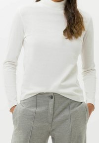 Frau trägt einen weißen Langarmrollkragenpullover und hochgeschnittene hellgraue Hose mit den Händen in den Taschen, lässige Pose.