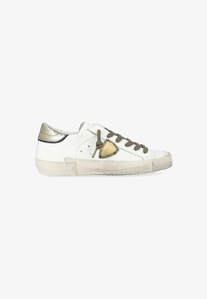 Des baskets blanches en cuir avec un talon doré métallique, des lacets gris texturés et un patch triangulaire doré sur le côté. Semelle usée visible.