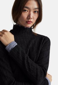 Maglione nero a collo alto con finitura texturizzata, indossato sopra una maglietta grigia chiara a maniche lunghe. Collo alto, design aderente.