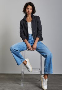 Musta vetoketjupusero, valkoinen T-paita, vaaleansiniset leveälahkeiset farkut ja valkoiset tennarit. Malli istuu kirkkaalla akryylijakkaralla. Neutraali tausta.