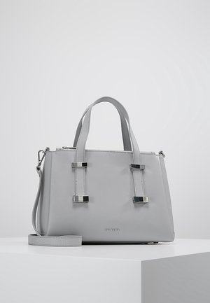 Sac à main en cuir gris clair avec deux poignées supérieures, accents métalliques argentés, bandoulière amovible et logo Ted Baker sur le devant, posé sur une surface blanche.