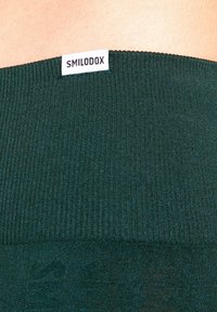 Tessuto verde scuro a coste con un motivo a maglia orizzontale nella parte superiore. Etichetta bianca con la scritta "SMILODOX" appare al bordo superiore.