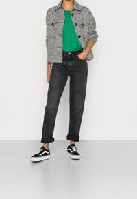 Grå texturerad jacka, grön t-shirt, svarta högmidjade jeans med uppvikt kant, svart- och vitfärgade sneakers, minimalistisk bakgrund.