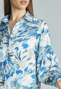 Blusa floral en vibrantes tonos azules y verdes, con cuello tipo solapa, cierre de botones y mangas abullonadas hechas de una tela suave.