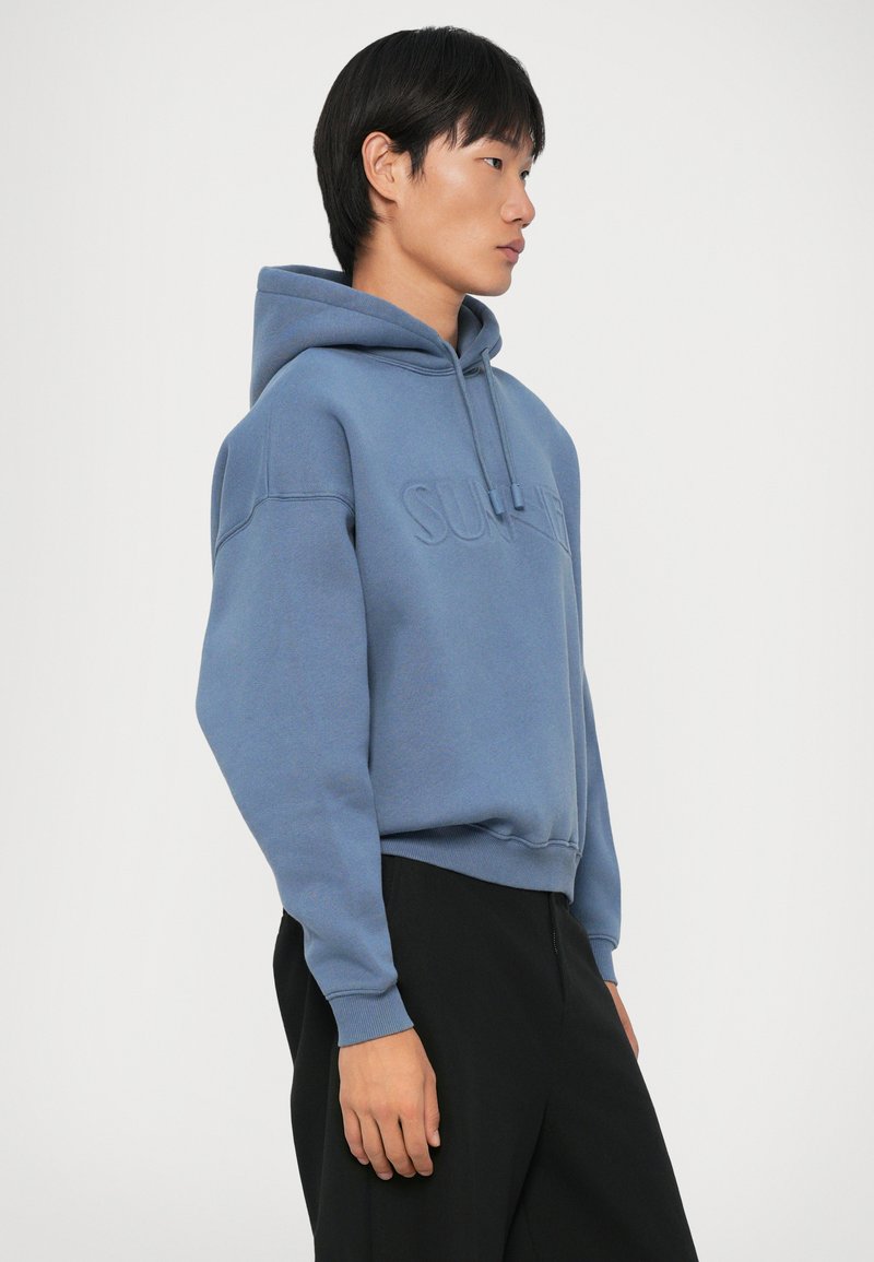 Felpa azzurra chiara con logo in rilievo sul davanti, maniche lunghe, cappuccio con cordoncino e polsini a coste. Indossata con pantaloni neri.