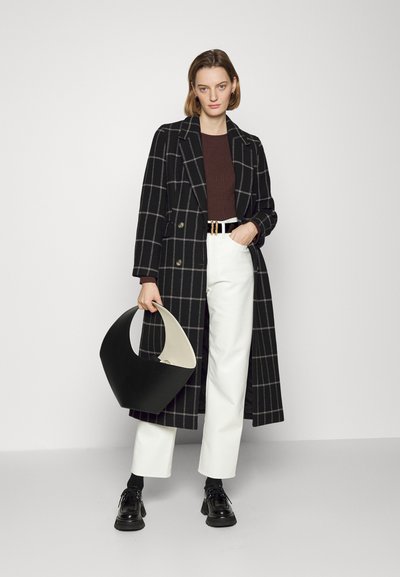 Lungo cappotto a quadri bianco e nero su un top marrone a coste, pantaloni bianchi e scarpe nere, che tiene una borsa curva bianca e nera.