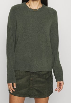 Femme portant un pull vert foncé à manches longues avec des poignets côtelés et une jupe en velours côtelé vert foncé à boutons.