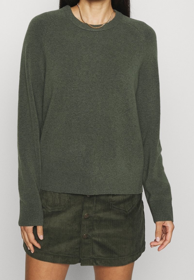 Femme portant un pull vert foncé à manches longues avec des poignets côtelés et une jupe en velours côtelé vert foncé à boutons.