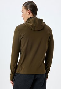 Chaqueta marrón con capucha, de manga larga y diseño ajustado, fabricada con un tejido suave y elástico. Presenta costuras visibles y un corte casual.