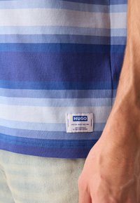 Camiseta a rayas azul hecha de algodón. Presenta diferentes tonos de rayas azules, un cuello redondeado y una etiqueta blanca con texto azul en el dobladillo.