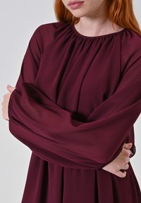 Donna con capelli rossi che indossa un vestito lungo e ampio a maniche lunghe di color bordeaux con scollatura arricciata, con le braccia incrociate davanti a uno sfondo chiaro e semplice.