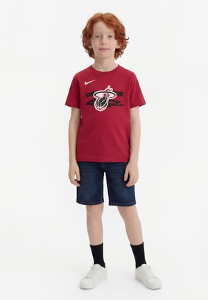 Garçon aux cheveux roux portant un t-shirt rouge Miami Heat, un short en jean foncé, des chaussettes noires et des baskets blanches, debout devant un fond blanc.