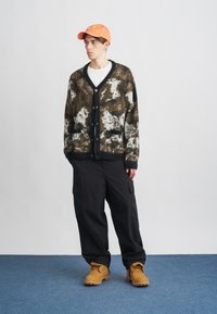 Smeđi i kremasti kardigan s uzorkom, crni detalji i gumbe sprijeda; uparen s crnim cargo hlačama i narančastom kapom, završeno s bež čizmama.