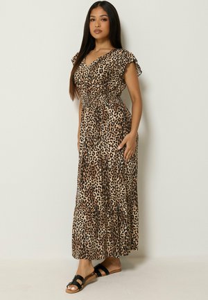 Mujer con cabello largo y oscuro que lleva un vestido largo sin mangas con estampado de leopardo y sandalias negras, de pie frente a un fondo claro y liso.