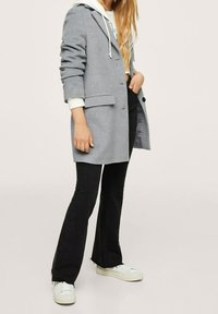 Šedý vlněný kabát s klopami a bočními kapsami, vrstvený přes bílou mikinu, spárovaný s černými zvonovými kalhotami a bílými teniskami.