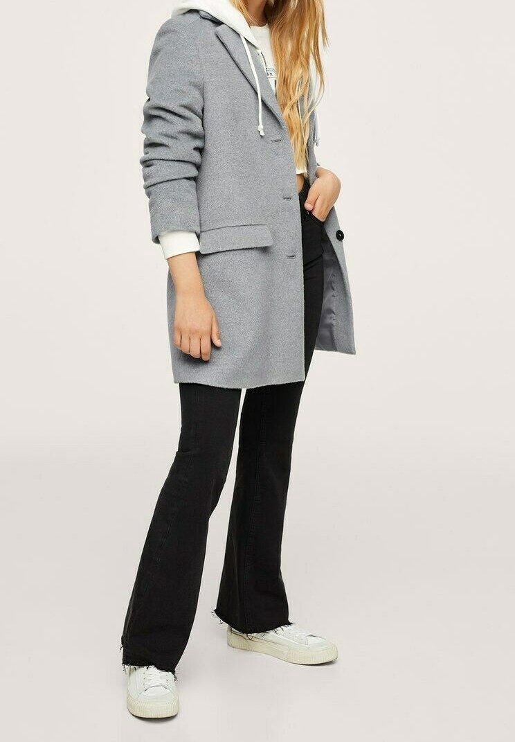 Šedý vlněný kabát s klopami a bočními kapsami, vrstvený přes bílou mikinu, spárovaný s černými zvonovými kalhotami a bílými teniskami.