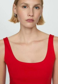 Mladá žena s krátkými blond vlasy, která má na sobě stříbrné kruhové náušnice a červený top bez rukávů, na neutrálním pozadí.