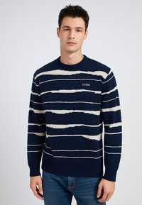 Maglione lavorato a maglia blu navy con strisce orizzontali bianche, collo a barcos, polsini e orlo a coste; presenta il logo "GUESS" sul davanti.