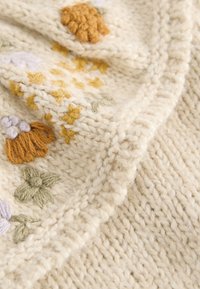 Knit blanket in creamy beige with textured yarn. Features colorful embroidery of flowers in yellow, purple, and green, creating a cozy appearance.