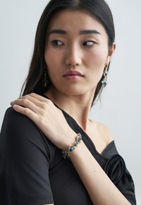 Armband met blauwe edelstenen, gouden accenten en bloemmotief. Draagt aan de pols met een zwarte off-shoulder top. Oorbellen passen bij het ontwerp van de armband.