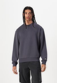 Mörkgrå sweatshirt med rundad halsringning, sänkt axelparti och ribbade mudd. Har en liten logotyp vid fållen och en avslappnad passform.