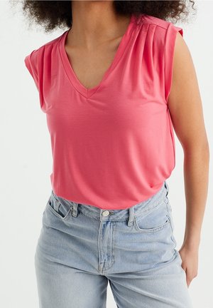 T-shirt z dekoltem w serek i krótkimi rękawami w jasnoróżowym kolorze, z subtelnymi pliskami na ramionach, zestawiony z jasnoniebieskimi dżinsami o wysokim stanie. Miękka tkanina, swobodny krój.