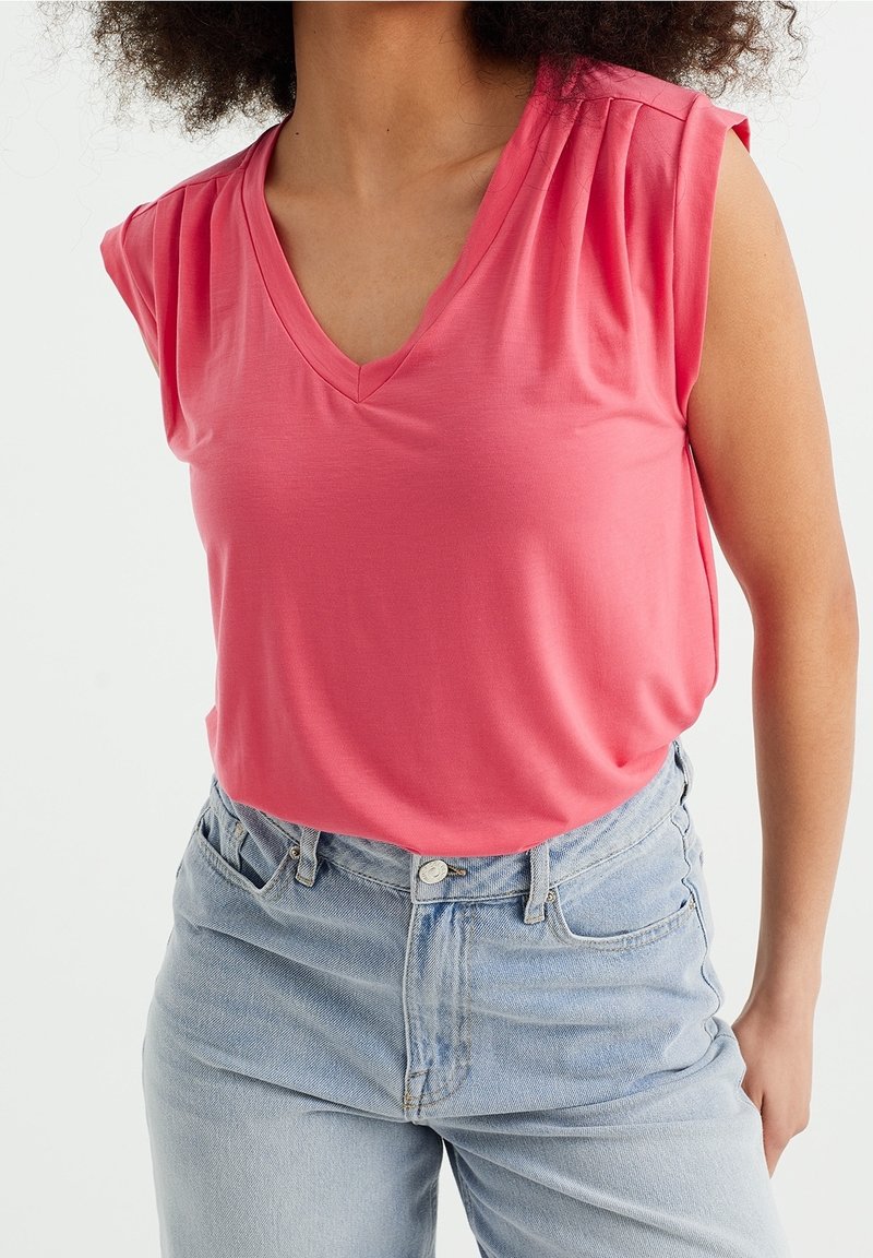 Halenka s výstřihem do V a krátkým rukávem v jasně růžové barvě se subtilními plisé na ramenech, spárovaná s světle modrými džíny s vysokým pasem. Měkká textura, ležérní design.
