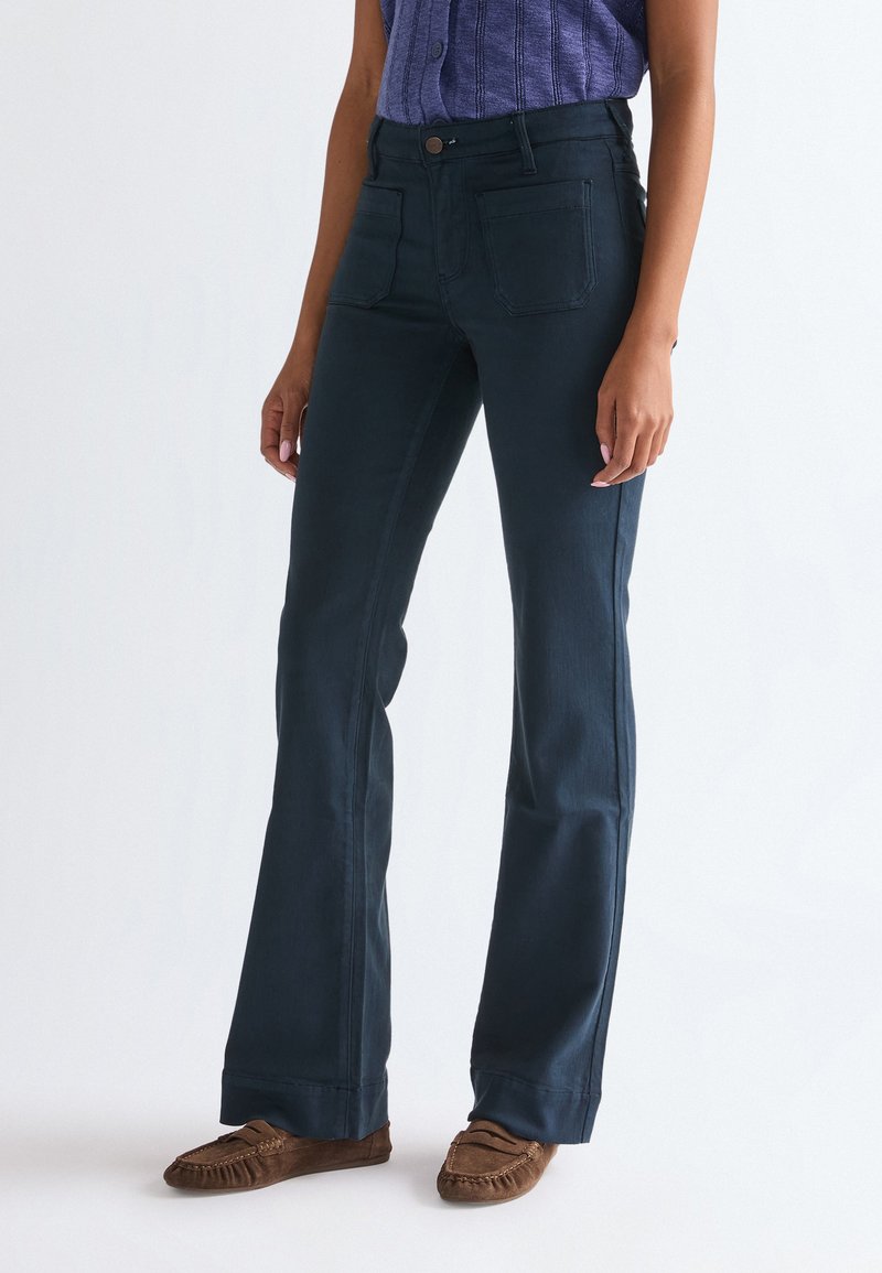 Femme portant un pantalon évasé bleu foncé, des mocassins marron et un haut violet sans manches en côtes, debout devant un fond blanc uni.