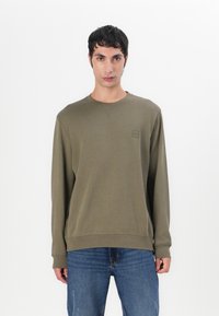 Olivengrønn genser med lange ermer, ribbestrikkede mansjetter og et subtilt logo-patch. Kombinert med blå slitte jeans for en avslappet stil.
