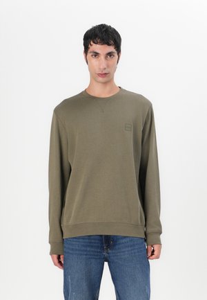 Olive green sweatshirt with long sleeves, ribbed cuffs, and a subtle logo patch. Paired with blue distressed jeans for a relaxed fit.
