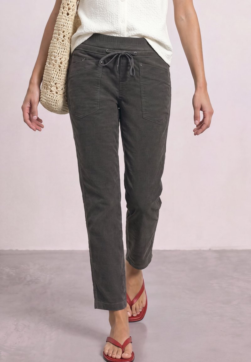 Woman wearing dark drawstring pants, white sleeveless top, red sandals, and carrying a beige crochet bag walking on a light floor.