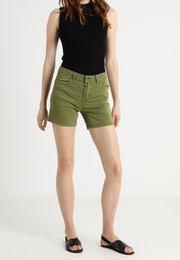 Pantalones cortos de mezclilla verde con un diseño clásico de cinco bolsillos y cierre de botón, combinados con una blusa sin mangas negra y sandalias planas negras.