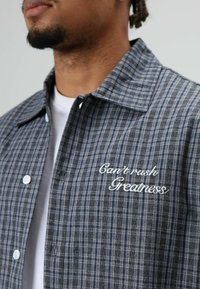 Grijs geruit shirt met knopen en witte knopen, met geborduurde tekst "Can't rush Greatness" aan de linkerkant, gladde stoftextuur.