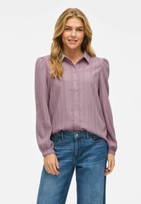 Langermet, lys lilla bluse med knappfront, teksturert stoff, plisserte skuldre og dekorative utskjæringer langs sømmene. Kombinert med denimjeans.