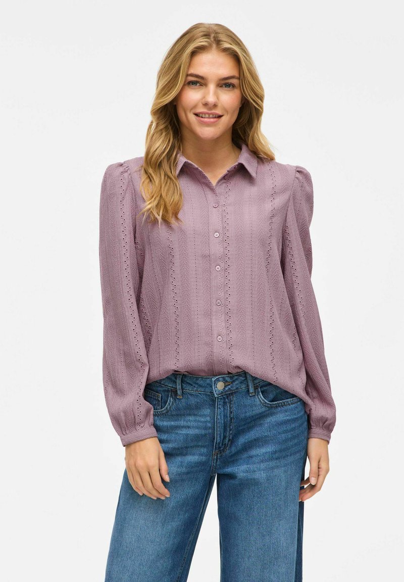 Langermet, lys lilla bluse med knappfront, teksturert stoff, plisserte skuldre og dekorative utskjæringer langs sømmene. Kombinert med denimjeans.