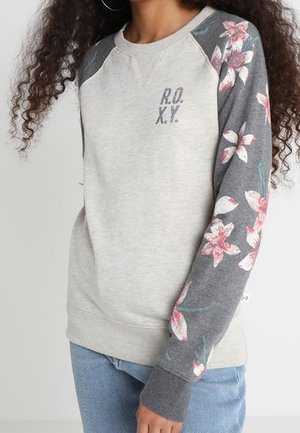 Femme portant un sweat-shirt gris clair avec des manches à motif floral gris foncé et le texte "R.O.X.Y." sur la poitrine, assorti à un jean bleu.