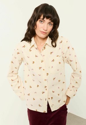 Frau mit dunklem welligem Haar, die ein cremefarbenes Hemd mit kleinen Blumenmustern und dunkelroten Hosen trägt, steht mit den Händen in der Hüfte.