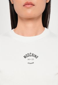 Balta medvilnės marškinėliai su juodu apskritimo logotipu, kuriame yra užrašas "MOSCHINO 86 23 JEANS". Apskritas iškirpimas ir trumpomis rankovėmis. Lygus audinys.