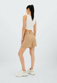 Shorts beiges avec des évents sur les côtés, associés à un débardeur court blanc. Le mannequin porte des baskets blanches. L'arrière-plan est d'un gris clair uni.