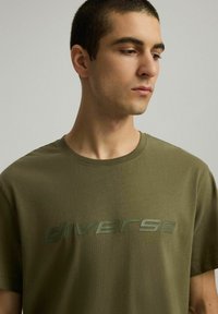 Oliwkowa bawełniana koszulka, z okrągłym dekoltem, z wypukłym, błyszczącym logo "diverse" w ciemniejszym zielonym kolorze na przodzie. Gładka faktura, krótkie rękawy.